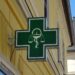 Insegna verde di una farmacia con il simbolo del mortaio e del serpente, su una parete gialla d'angolo.