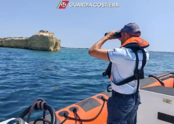 Militare della Guardia Costiera osserva il mare con il binocolo a bordo di un gommone, vicino a una costa rocciosa