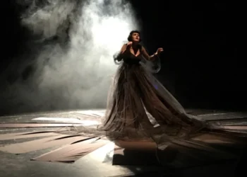 Donna in abito nero durante una performance teatrale sul palco, con fumo scenico e luci.