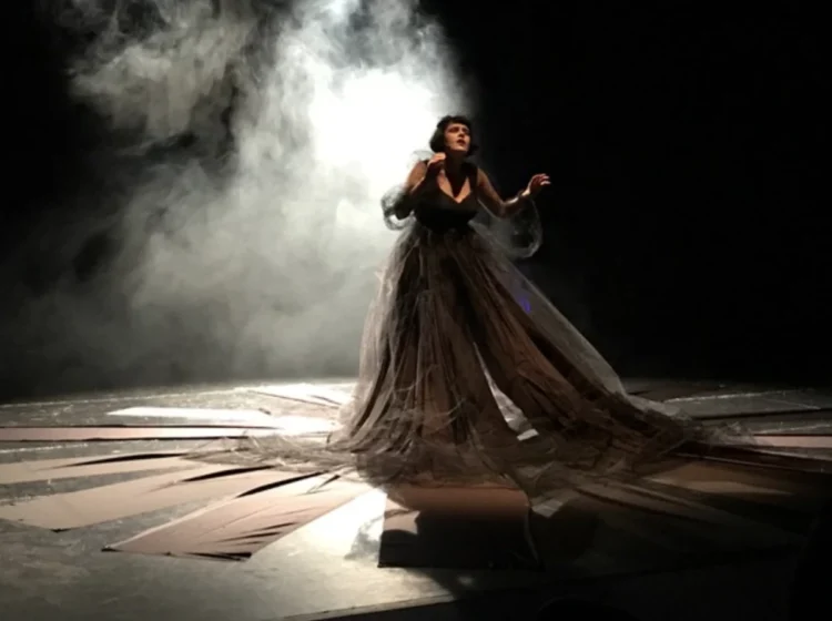 Donna in abito nero durante una performance teatrale sul palco, con fumo scenico e luci.