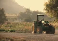 Trattore verde parcheggiato su sterrato in un'area agricola, con campi coltivati e colline sullo sfondo al tramonto.