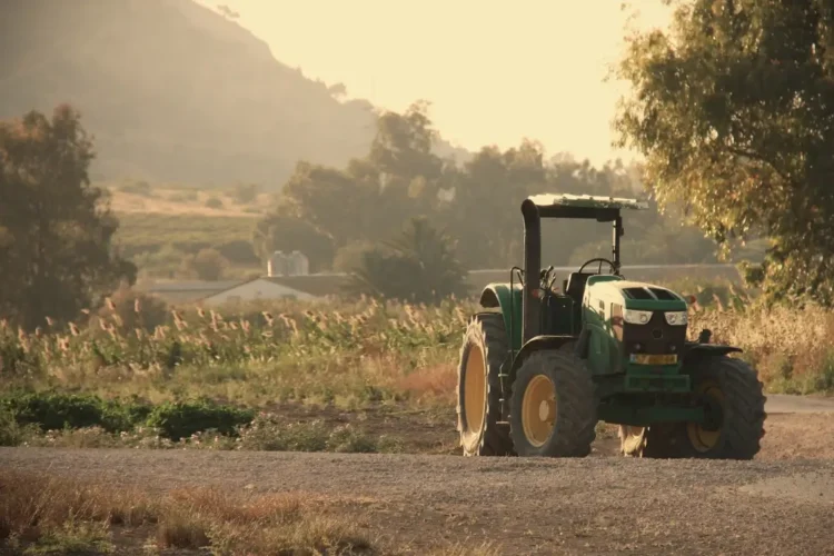 Trattore verde parcheggiato su sterrato in un'area agricola, con campi coltivati e colline sullo sfondo al tramonto.