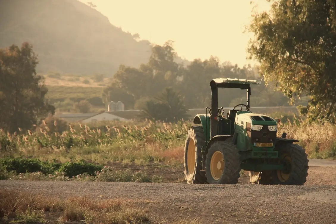 Trattore verde parcheggiato su sterrato in un'area agricola, con campi coltivati e colline sullo sfondo al tramonto.