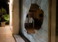 Vetrata di una casa sfondata durante la notte, con vetri rotti sul pavimento e interno dell'abitazione visibile.