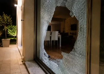 Vetrata di una casa sfondata durante la notte, con vetri rotti sul pavimento e interno dell'abitazione visibile.