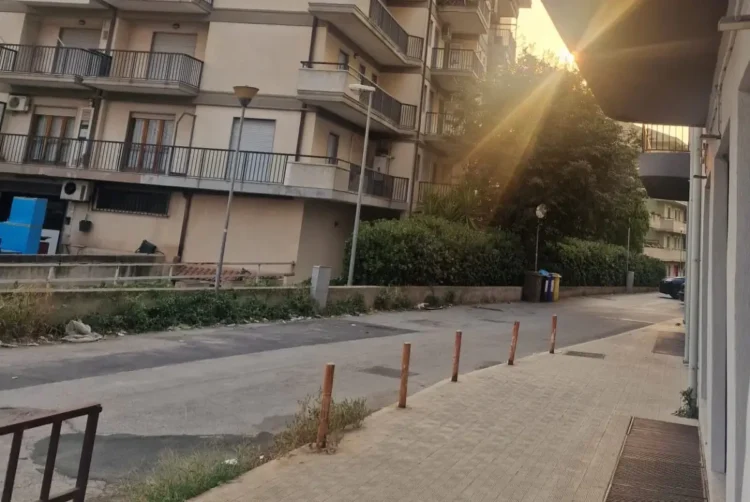 Una via residenziale di Modica con marciapiede, dissuasori e cassonetti per la raccolta differenziata, al tramonto