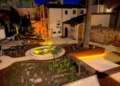 Giardino con fontana circolare e piante mediterranee nella piazzetta illuminata di notte, tra edifici storici in pietra