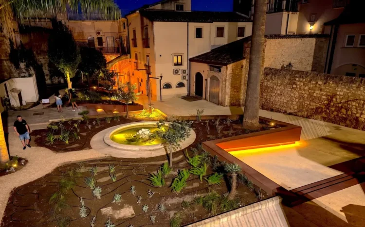 Giardino con fontana circolare e piante mediterranee nella piazzetta illuminata di notte, tra edifici storici in pietra
