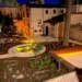 Giardino con fontana circolare e piante mediterranee nella piazzetta illuminata di notte, tra edifici storici in pietra