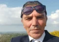 Giuseppe Damasco in un primo piano, giacca scura e camicia bianca, sullo sfondo cielo e vegetazione.