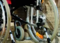 Primo piano delle ruote e del telaio di una sedia a rotelle su fondo ghiaioso, con struttura in vimini sullo sfondo