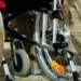 Primo piano delle ruote e del telaio di una sedia a rotelle su fondo ghiaioso, con struttura in vimini sullo sfondo