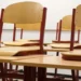 Banchi e sedie di legno vuoti in un'aula scolastica, ripresi in prospettiva con luce naturale.