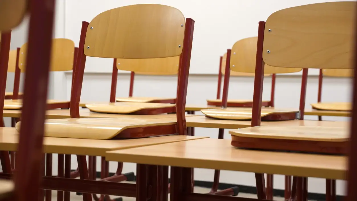Banchi e sedie di legno vuoti in un'aula scolastica, ripresi in prospettiva con luce naturale.