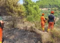 Squadre antincendio in tuta arancione osservano un'area boschiva bruciata su un versante collinare
