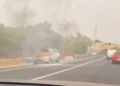 Suv bianco in fiamme a bordo strada, con fumo nero e una persona vicino al veicolo in fiamme