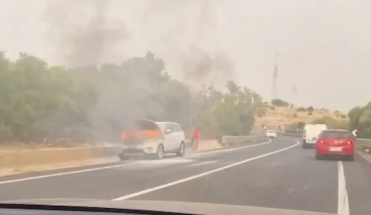 Suv bianco in fiamme a bordo strada, con fumo nero e una persona vicino al veicolo in fiamme
