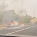 Suv bianco in fiamme a bordo strada, con fumo nero e una persona vicino al veicolo in fiamme