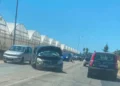 Auto scura con cofano aperto e frontale danneggiato ferma sulla provinciale, coda di veicoli e serre sullo sfondo.