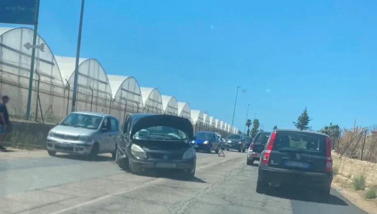 Auto scura con cofano aperto e frontale danneggiato ferma sulla provinciale, coda di veicoli e serre sullo sfondo.