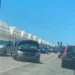 Auto scura con cofano aperto e frontale danneggiato ferma sulla provinciale, coda di veicoli e serre sullo sfondo.