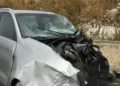 Auto di colore argento con la parte anteriore completamente distrutta e il parabrezza in frantumi, ferma sul ciglio della strada