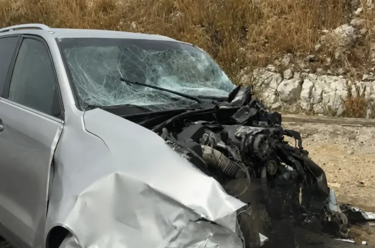 Auto di colore argento con la parte anteriore completamente distrutta e il parabrezza in frantumi, ferma sul ciglio della strada