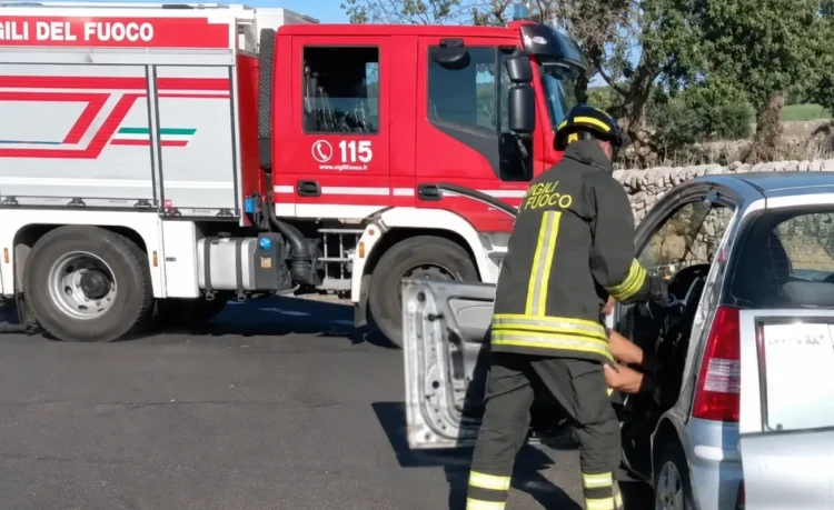 Vigile del fuoco in tenuta da intervento estrae uno sportello da un'auto incidentata, autobotte dei vigili del fuoco sullo sfondo.