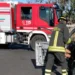 Vigile del fuoco in tenuta da intervento estrae uno sportello da un'auto incidentata, autobotte dei vigili del fuoco sullo sfondo.