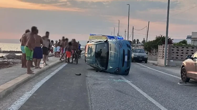 Fiat 600 rovesciata sul fianco sulla SP67 tra Marza e Pozzallo, con ambulanza e passanti sullo sfondo al tramonto