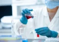 Mani con guanti blu versano un liquido rosso in una provetta tra beute e provette di laboratorio