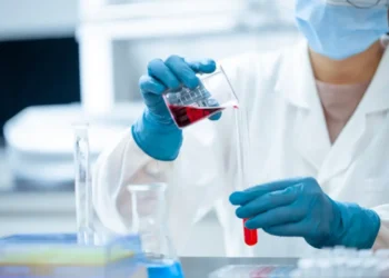 Mani con guanti blu versano un liquido rosso in una provetta tra beute e provette di laboratorio