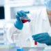 Mani con guanti blu versano un liquido rosso in una provetta tra beute e provette di laboratorio