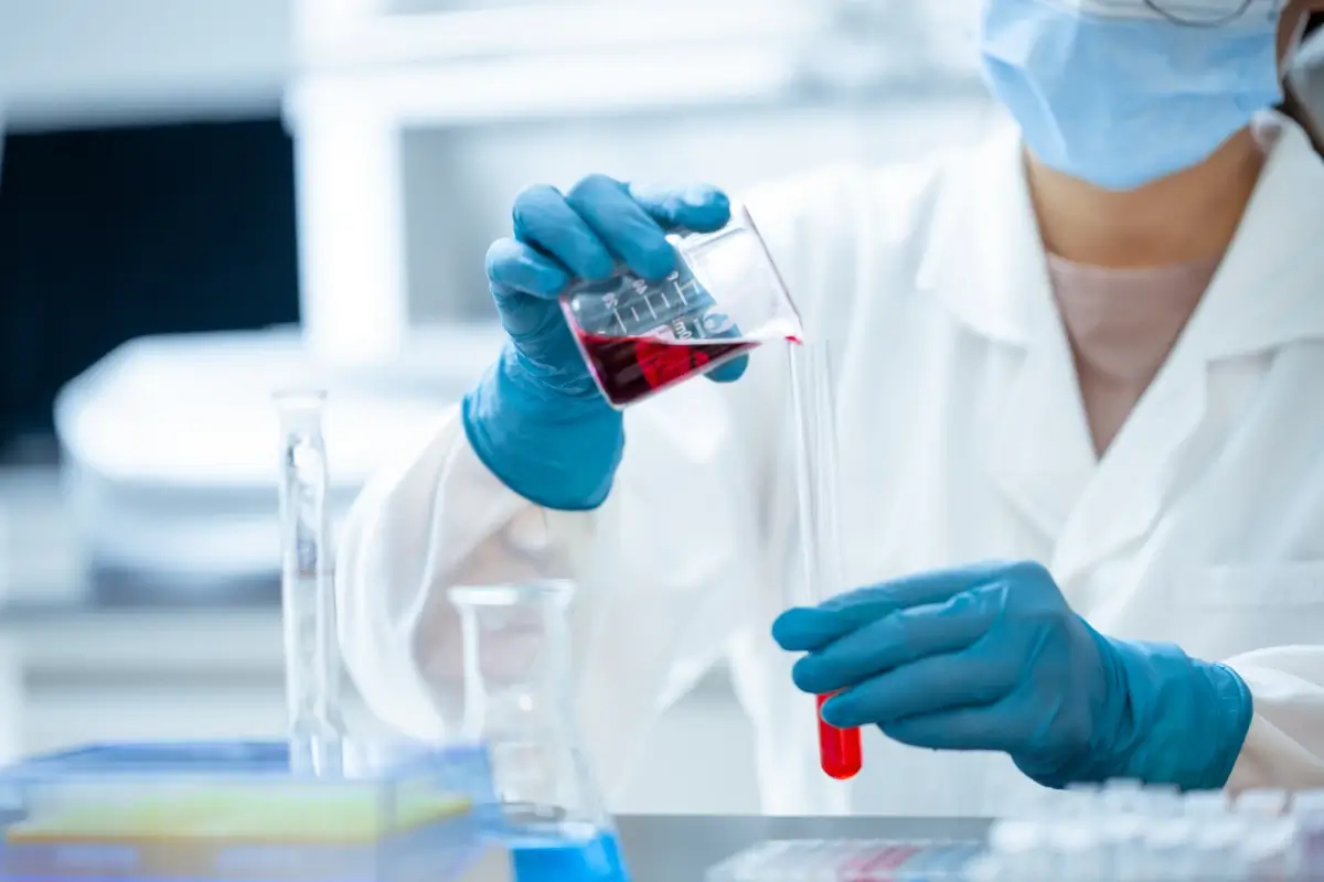 Mani con guanti blu versano un liquido rosso in una provetta tra beute e provette di laboratorio