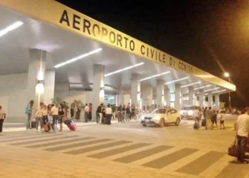 Ingresso dell'aeroporto civile di Comiso di sera, con viaggiatori e valigie davanti al terminal illuminato.