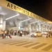 Ingresso dell'aeroporto civile di Comiso di sera, con viaggiatori e valigie davanti al terminal illuminato.
