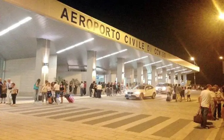 Ingresso dell'aeroporto civile di Comiso di sera, con viaggiatori e valigie davanti al terminal illuminato.