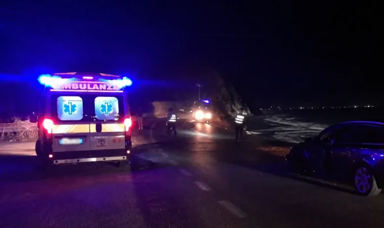 Ambulanza e auto della polizia su una strada costiera di notte, accanto a un'auto incidentata con la portiera aperta.