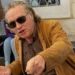 Uomo anziano con capelli grigi, occhiali da sole scuri e giacca ocra seduto a un tavolino da bar all'aperto.