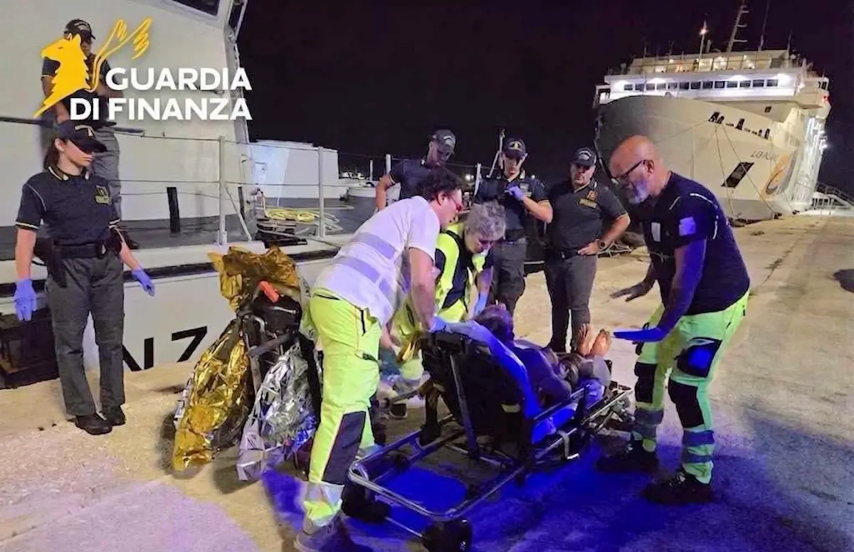 Soccorritori e Guardia di Finanza assistono un uomo su una barella in banchina di notte, traghetto sullo sfondo.