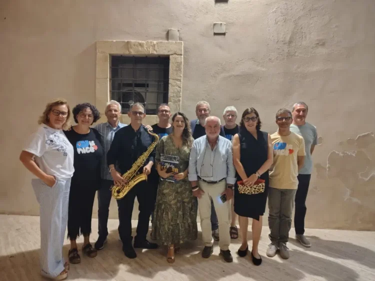 Gruppo di circa dodici persone in posa all'aperto di notte, uno dei quali regge un sassofono dorato, davanti a un muro in pietra