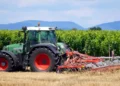Trattore verde con attrezzatura agricola in un vigneto, campo di stoppie in primo piano e colline sullo sfondo.
