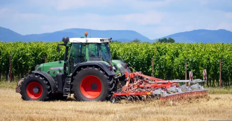 Trattore verde con attrezzatura agricola in un vigneto, campo di stoppie in primo piano e colline sullo sfondo.