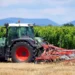 Trattore verde con attrezzatura agricola in un vigneto, campo di stoppie in primo piano e colline sullo sfondo.