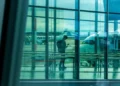 Passeggero con zaino consulta lo smartphone nell'area imbarchi di un aeroporto, aeromobili parcheggiati sul piazzale sullo sfondo.