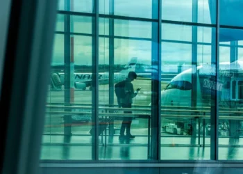 Passeggero con zaino consulta lo smartphone nell'area imbarchi di un aeroporto, aeromobili parcheggiati sul piazzale sullo sfondo.