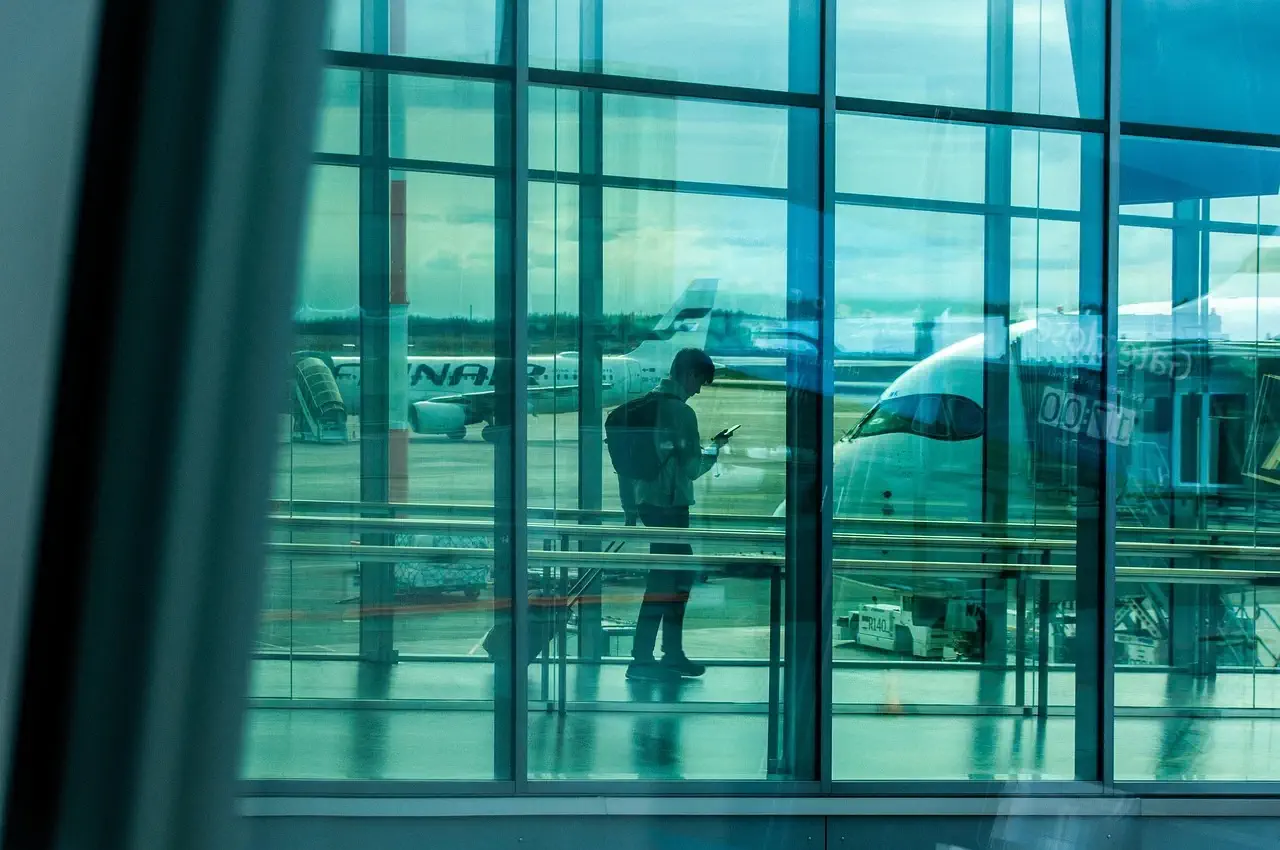 Passeggero con zaino consulta lo smartphone nell'area imbarchi di un aeroporto, aeromobili parcheggiati sul piazzale sullo sfondo.