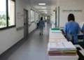 Corridoio di ospedale di comunità con infermiere in divisa blu al bancone e cartelle cliniche impilate in primo piano