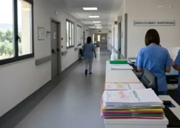 Corridoio di ospedale di comunità con infermiere in divisa blu al bancone e cartelle cliniche impilate in primo piano