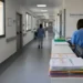 Corridoio di ospedale di comunità con infermiere in divisa blu al bancone e cartelle cliniche impilate in primo piano